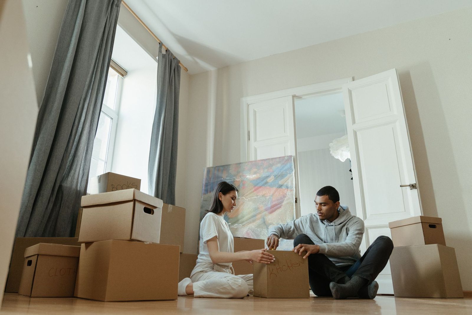 A couple sitting on the floor of an empty flat, unpacking moving boxes labelled books, clothes and kitchen